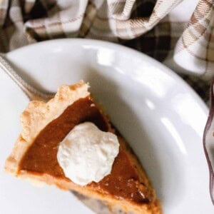 overhead photo of a piece of pumpkin pie on a cake knife being served onto a white platter with a corner of the pie in the pie plate to the right and a plaid towel in the background