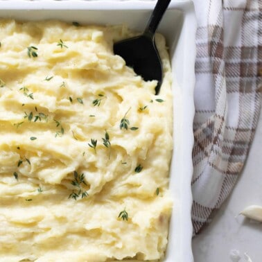 garlic mashed potatoes topped with herbs in a white baking dish with a black spoon in the potatoes. The baking dish rests on a tan and white plaid towel