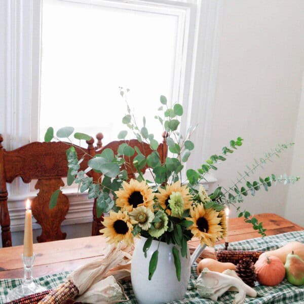 a vintage wood table with a green checked table runner topped with pumpkins, Indian corn, and a vase full of eucalyptus and sunflowers