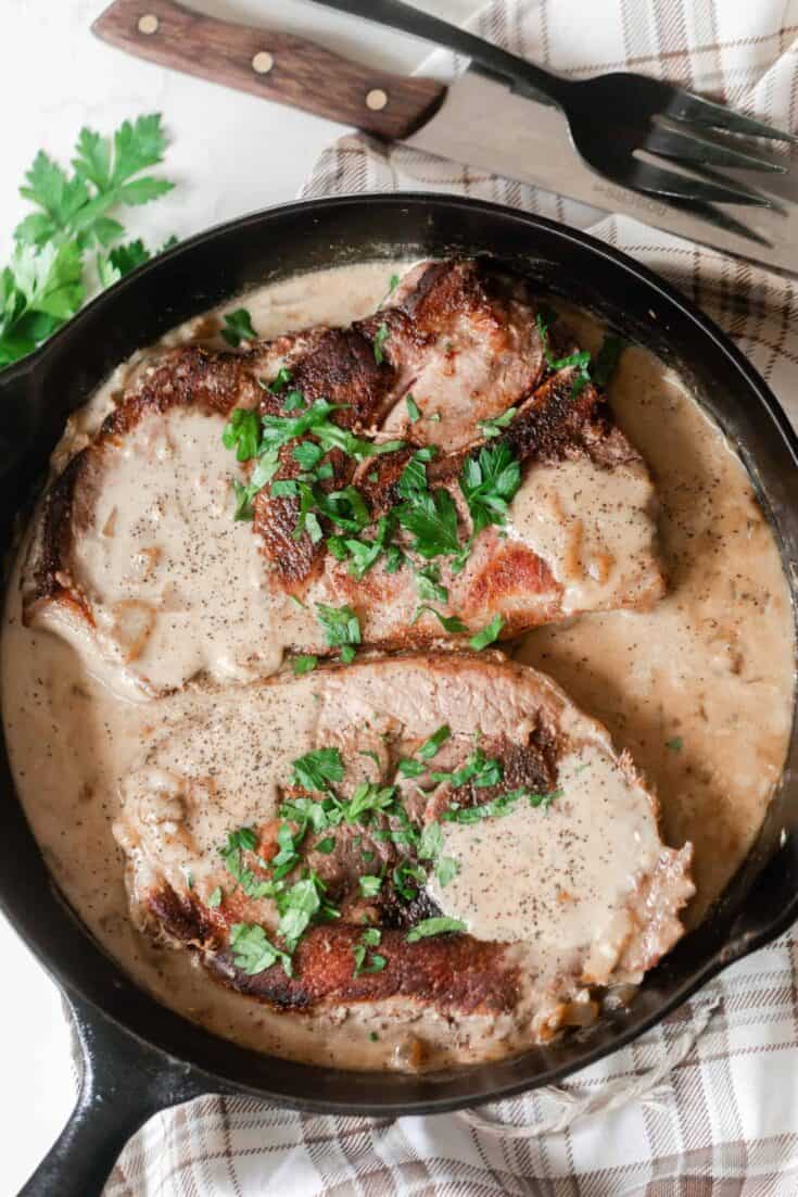 pork steaks in a creamy onion sauce and topped with fresh parsley in a cast iron skillet on a cream and brown plain towel with a fork and steak knife above the skillet