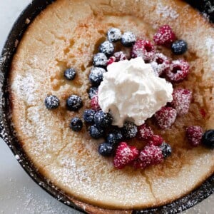 overhead photo of a cast iron skillet with a sourdough German pancake topped with berries, whipped cream, and powdered sugar