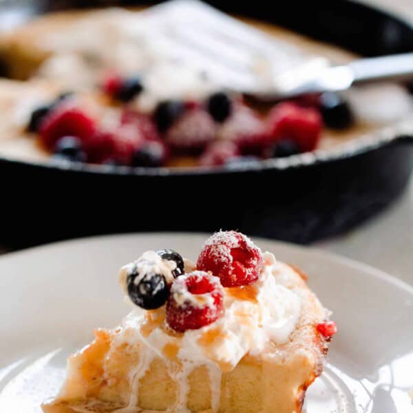 slice of sourdough dutch baby pancake topped with maple syrup, cream, and berries on a white plate with a cast iron skillet with more pancakes in the background