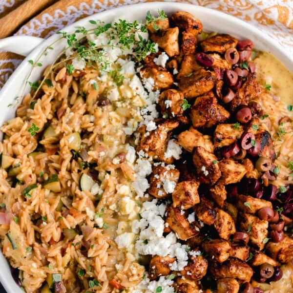 overhead photo of Greek chicken with orzo pasta, feta, and retables in a white enameled cast iron skillet on a gray countertop and a mustard colored towel to the left