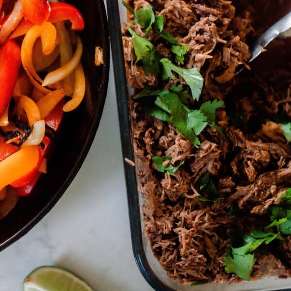 a glass dish of beef carnitas topped with cilantro. A cast iron skillet with peppers and onions are to the left and lime wedges are in front