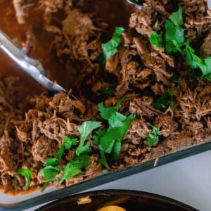 overhead photo of beef carnita meat topped with fresh cilantro in a glass backing dish with tongs and a cast iron skillet with cooked peppers and onions