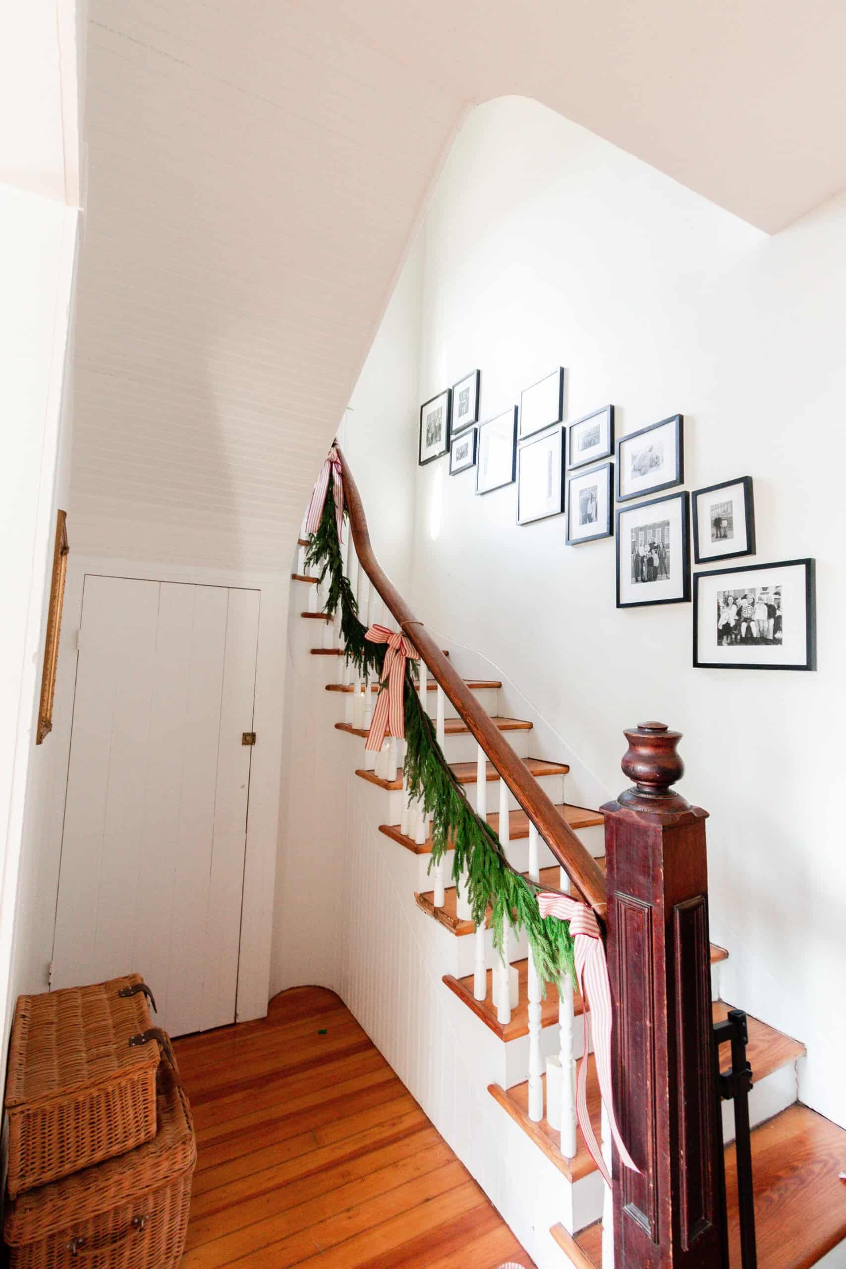 picture of a stairway wrapped in green garland and tied with ribbon. A gallery wall full of family pictures is going up the wall of the stairs