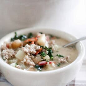 bowl of zuppa toscana soup with a spoon on a cream and brown plain napkin with a white dutch oven in the background