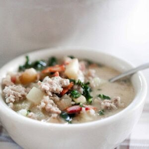 bowl of zuppa toscana soup with a spoon on a cream and brown plain napkin with a white dutch oven in the background