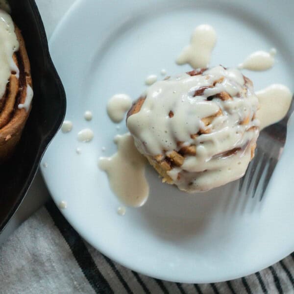 overhead photo of a einkorn cinnamon roll topped with cream cheese frosting on a white plate with a fork. A cast iron skillet with more rolls is behind the plate