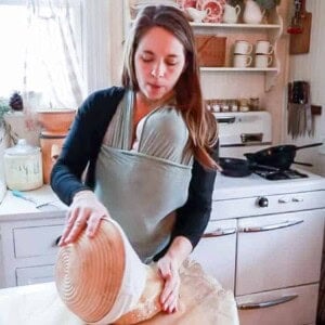 womann wearing a baby taking einkorn sourdough bread dough out of a banneton basket and placing it on a sheet of parchment paper.