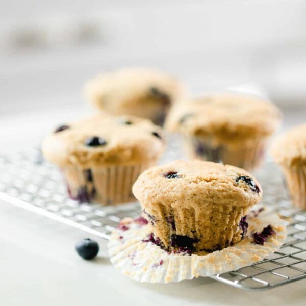 five sourdough discard blueberry muffins on a wire cooling rack on a white countertop with fresh blueberries sprinkled around. The muffin at the corner has the cupcake liner peeled down