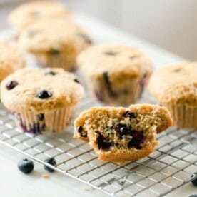 sourdough blueberry muffins on a wire rack on a white countertop with blueberries scattered around on the counter. The front muffin is sliced in half.