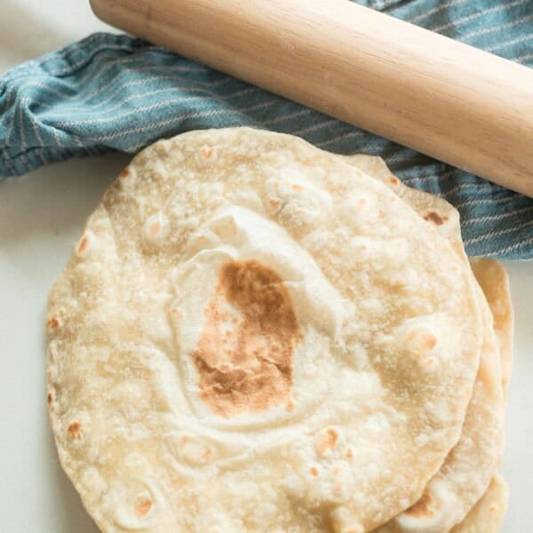 overhead photo of sourdough tortillas stack in a pile on a white quartz countertop with a blue stripped towel in the background with a rolling pin on top