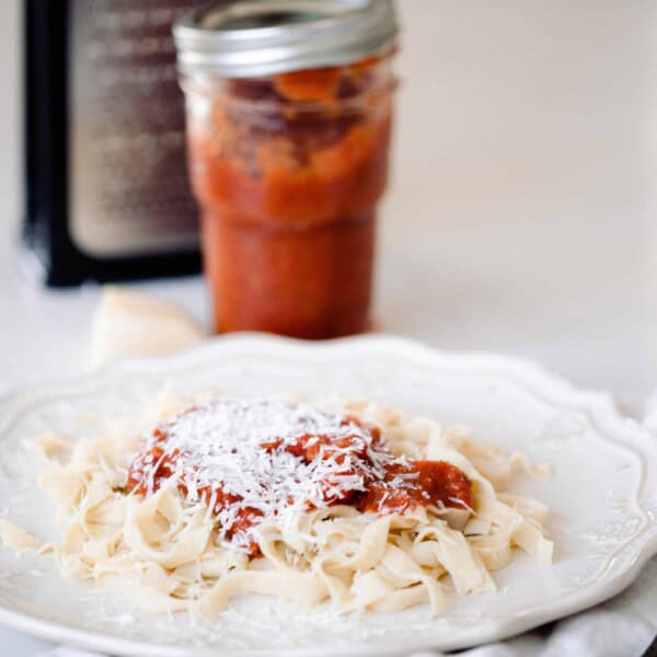 freshly cooked sourdough pasta topped with homemade marinara and grated parmesan cheese on a white fluted plate with a white and blue towel with a fork placed on top to the right. A jar of marina, cheese grater and a chunk of parmesan cheese are behind the plate.