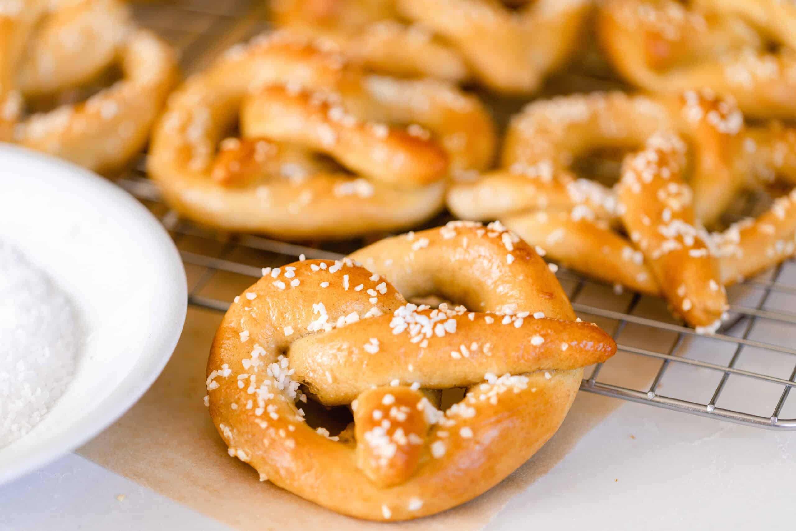 six sourdough pretzels layered on a counter and a wire cooling rack with a white bowl to the left.