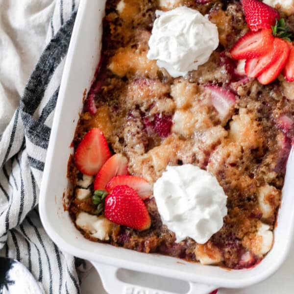 sourdough strawberry cobbler topped with fresh whipped cream and sliced strawberries. The cobbler is in a white baking dish on a white and black stripped towel