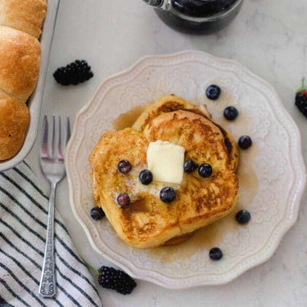 overhead picture of two brioche French toast slices stacked on top pf each other and topped with butter and fresh blueberries on a cream colored plate. The plate is on a white quartz countertop with a stripped towel and a loaf go brioche to the left