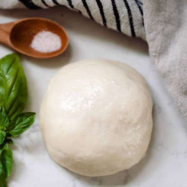 ball of homemade mozzarella cheese on a white countertop with a fresh sprig of basil, a wooden spoon with salt, and a black and white stripped towel surrounding the cheese