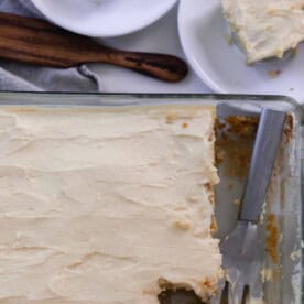 overhead photo of a baking dish with sourdough cake with a thick layer of buttercream frosting. A metal spatula is resting in the glass baking dish. Two square pieces of cake on white plates sit next to the baking dish.