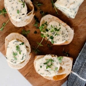 four slices of fresh bread with compound butter on top on a cutting board with herbs spread around and a log of herb butter to the back right