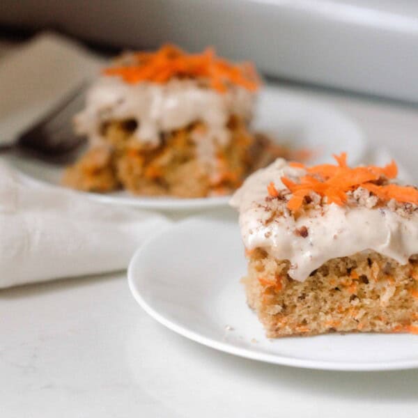 two slices of sourdough carrot cake topped with cream cheese frosting and a few shredded carrots and chopped pecans on white plates on a white countertop