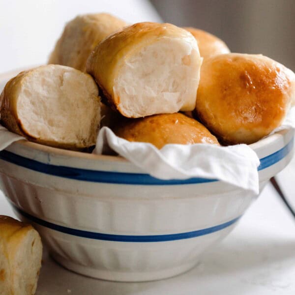 sourdough Hawaiian sweet rolls stacked in a white and cream ironstone bowl lined with a white linen towel. The bowl is on a white countertop