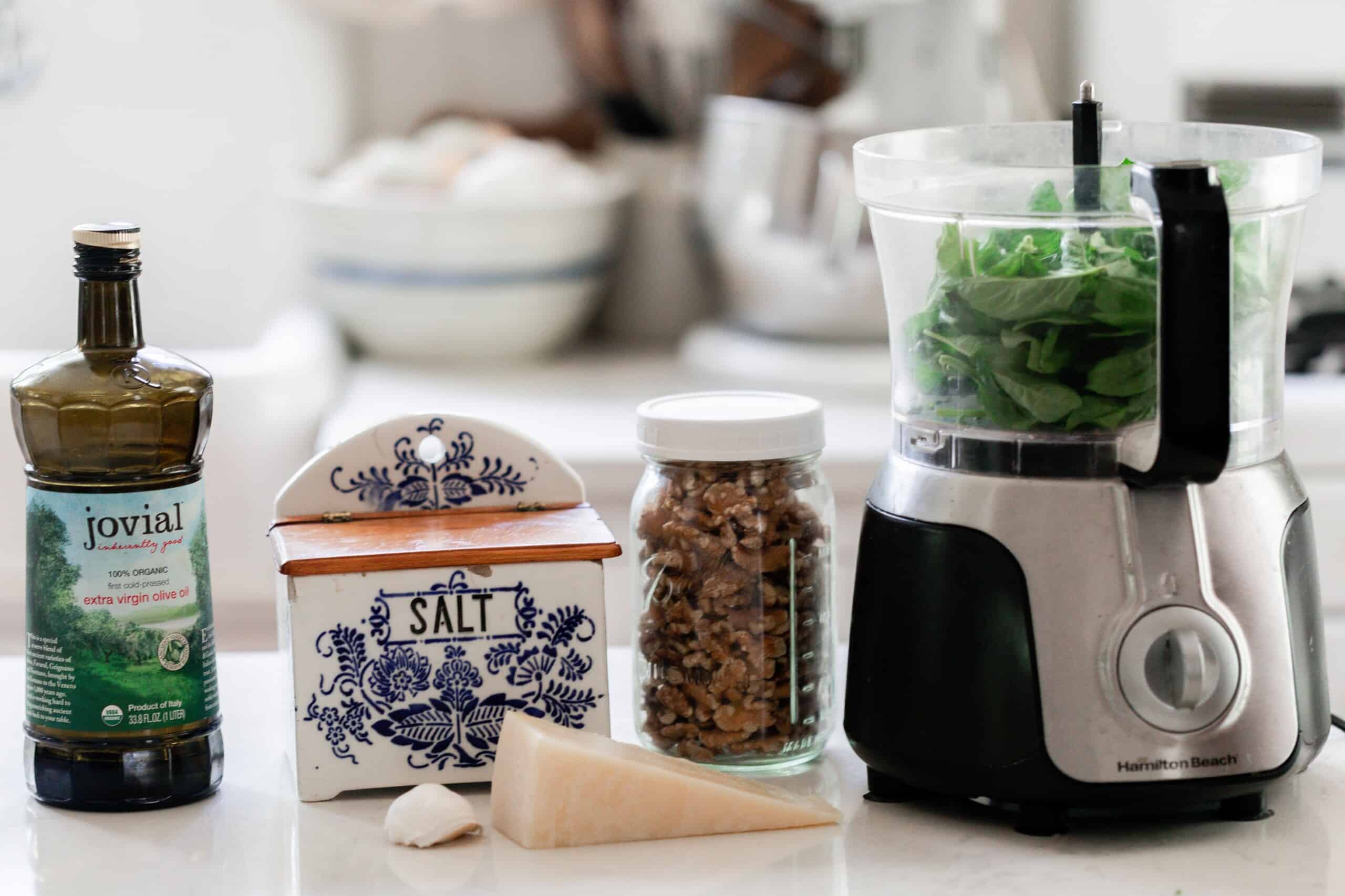 olive oil, salt, Parmesan, garlic, walnuts in a jar, and a food processor with basil on a white countertop in a kitchen.