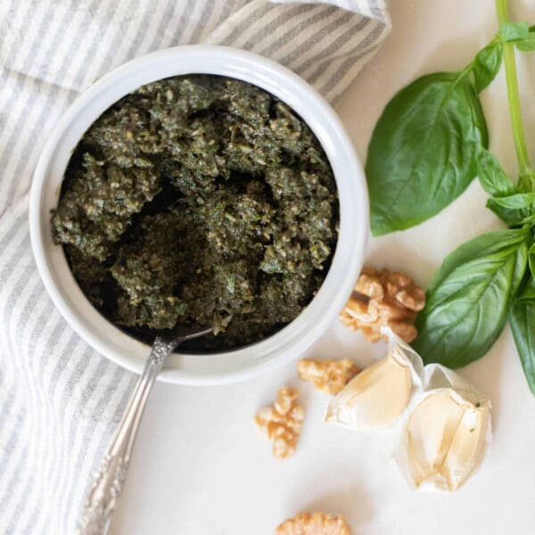 overhead photo of walnut pesto in a white jar with a spoon on a white countertop with walnuts and basil spread around the counter.