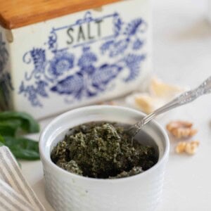 pesto in a white ramekin with a spoon on a white countertop with walnuts basil, a stripped towel and a vintage blue and white salt box in the background