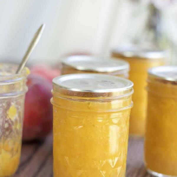 jars of canned peach preserves, including an opened jar with a knife, on a wood table. Peaches and flowers are in the background