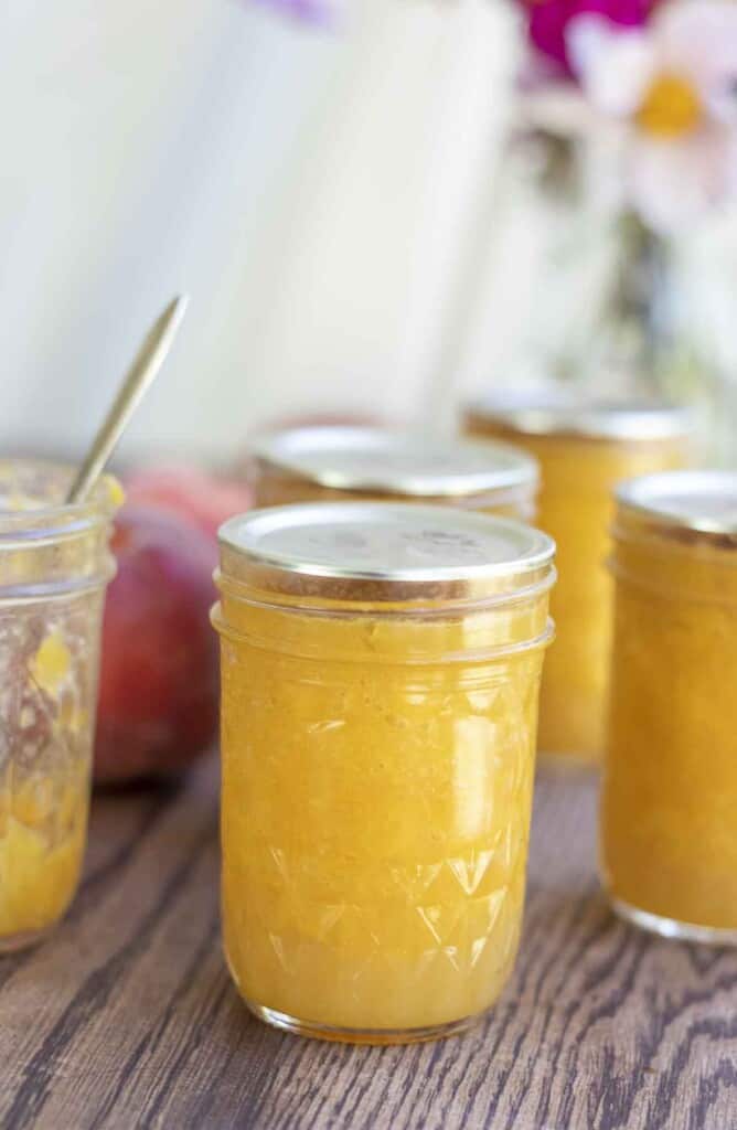 jars of canned peach preserves, including an opened jar with a knife, on a wood table. Peaches and flowers are in the background