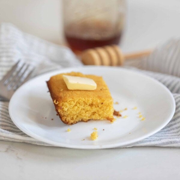 a slice of cornbread topped with butter on a white plate on a gray and white stripped towel on a white countertop. A Fork sits to the left and honey is in the background