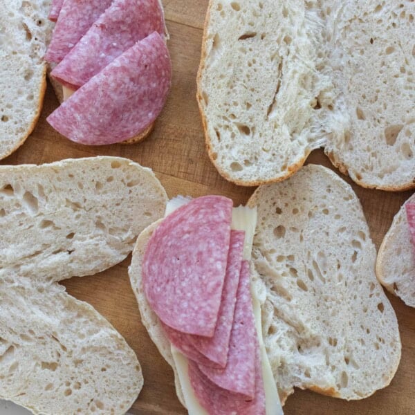 overhead photo of sourdough hoagie rolls sliced in half and topped with cheese and salami on a wood cutting board