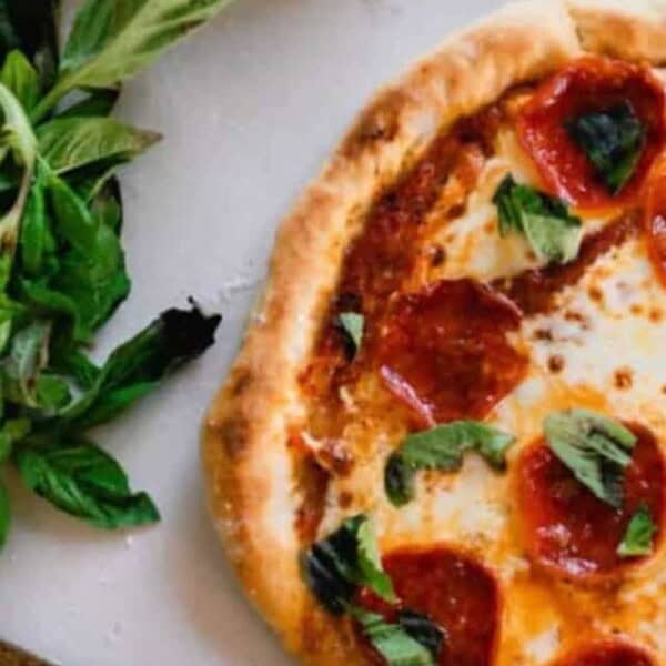 half of a sourdough pizza topped with sauce, cheese, pepperonis, and basil on a white countertop with fresh basil to the left