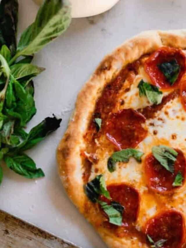 half of a sourdough pizza topped with sauce, cheese, pepperonis, and basil on a white countertop with fresh basil to the left