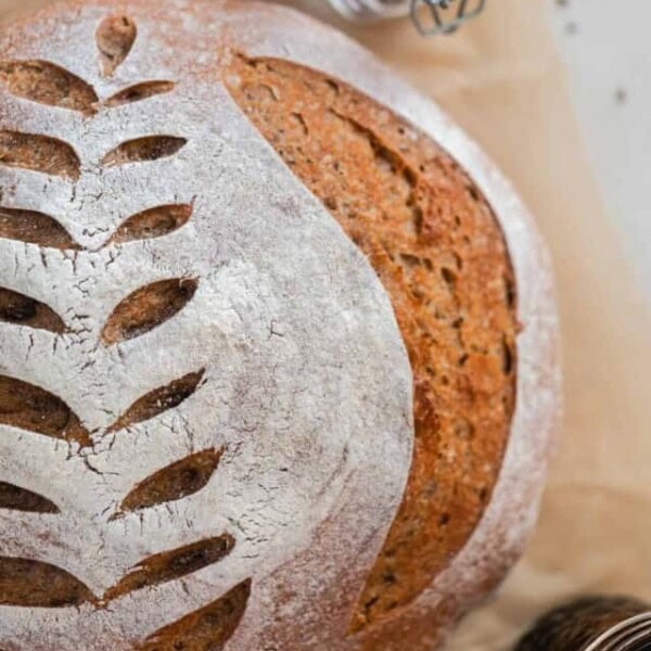 overhead photo of a crusty loaf of sourdough rye bread with a wheat design scored on top. The loaf rest on parchment paper with a jar of spices and a jar of molasses around the loaf