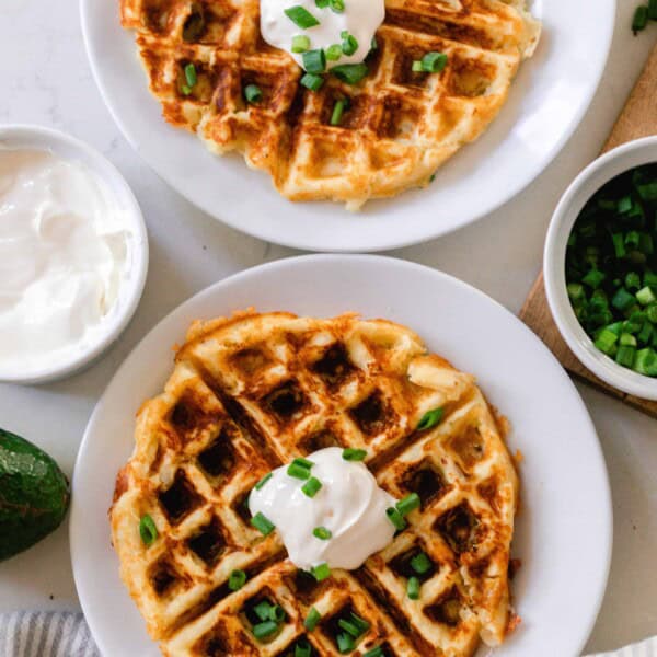 two potato waffles on a white plate topped with sour cream and chives. The plates rest on a white countertop with little jars of chives and sour cream