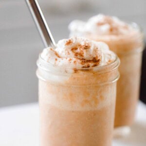 two pumpkin smoothies topped with whipped cream sprinkled with pumpkin spice. A metal straw is in the smoothie and the smoothies sit on a white countertop.