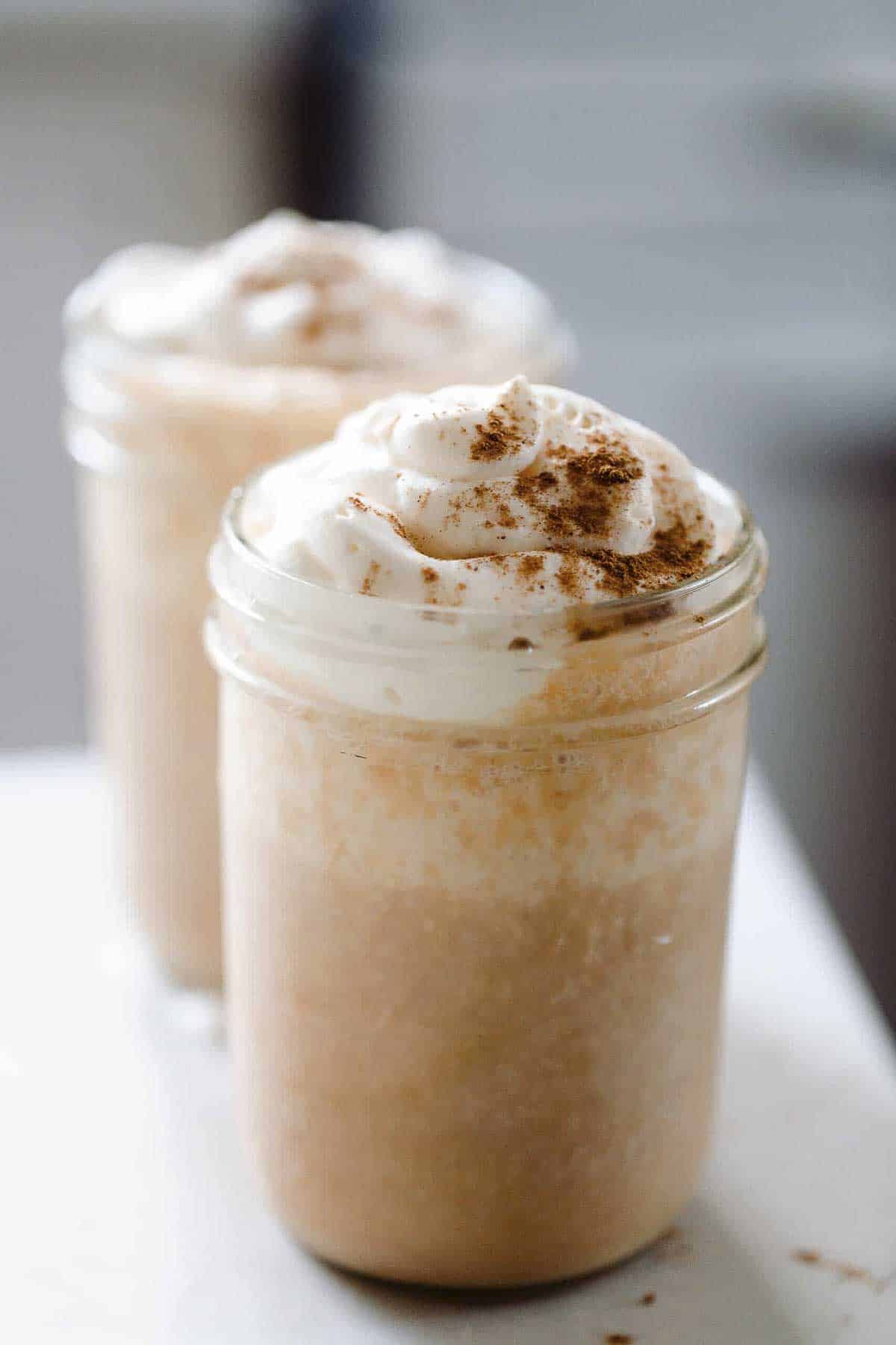 two pumpkin pie smoothies in glass mason jars topped with whipped cream and sprinkled with more pumpkin spice.