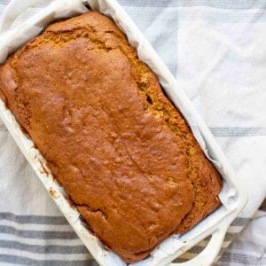 overhead photo of a loaf of sourdough pumpkin bread in a parchment lined stone bread pan on top of a blue and white stripped towel on a wood table