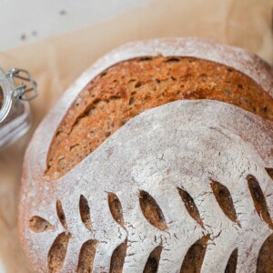 overhead photo of a crusty loaf of sourdough rye bread with a wheat design scored on top. The loaf rest on parchment paper with a jar of spices and a jar of molasses around the loaf