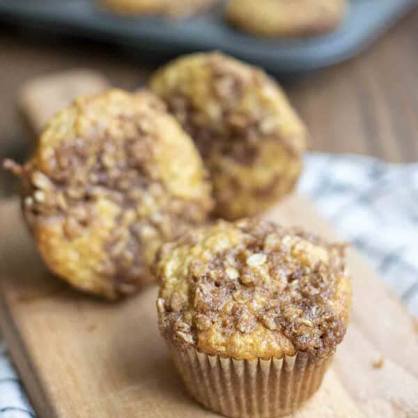 three sourdough banana muffin with a brown sugar oat crumble on a small wood cutting board with a tray of muffins in the background