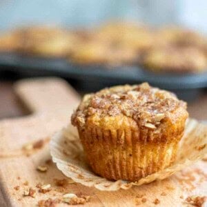 a unwrapped sourdough banana muffin with a oat crumble on a wood cutting board with oat crumble surrounding the muffin. There is a tray of muffins in the background