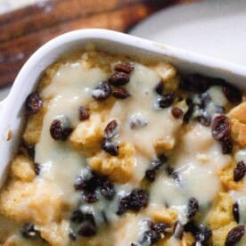 overhead photo of sourdough bread pudding topped with raisins and a creamy sauce in a white baking dish on a white countertop with a wooden spatula to the right