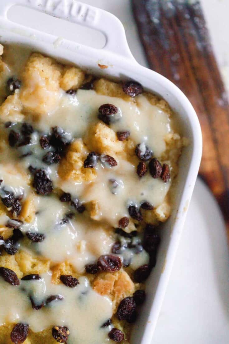 overhead photo of sourdough bread pudding topped with raisins and a creamy sauce in a white baking dish on a white countertop with a wooden spatula to the right