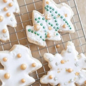 sourdough sugar cookies cut out as snowflakes and trees decorated with white frosting and gold balls. on a wire rack on parchment paper