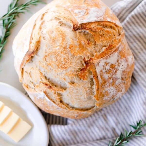 overhead photo of a loaf of crust rosemary sourdough bread on a gray and white towel. With fresh springs of rosemary around the loaf