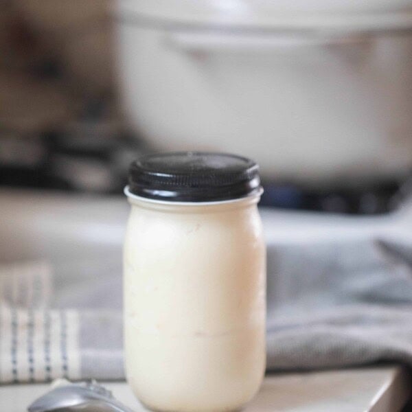 homemade lard in a glass mason jar with a black lid on a white countertop with a spoon in from of the jar. A white dutch oven is in the background