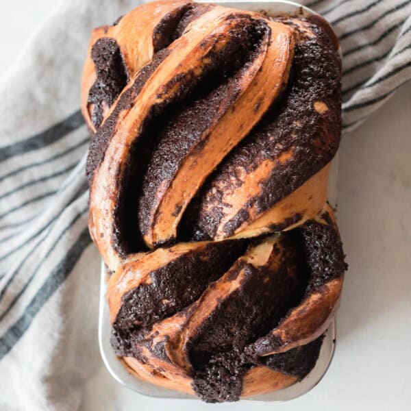 overhead photo of a loaf of sourdough chocolate babka on a white countertop and a black and cream colored stripped towel