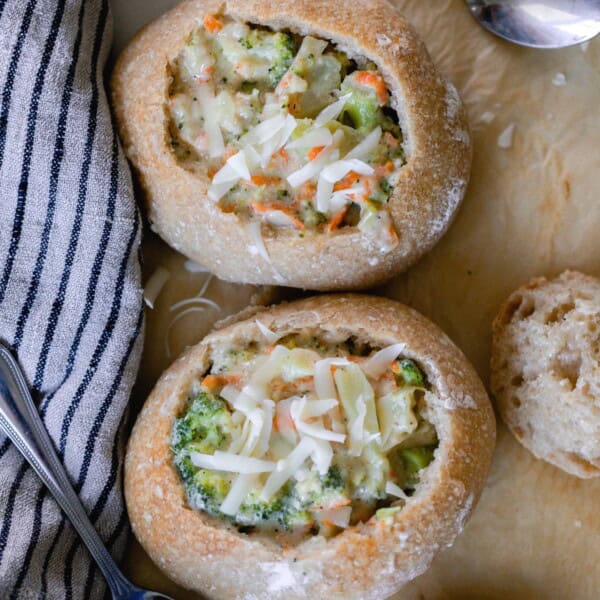 two sourdough bread bowls filled with broccoli cheddar soup and topped with cheese on parchment paper with spoons and a black and white stripped towel around the bread bowls